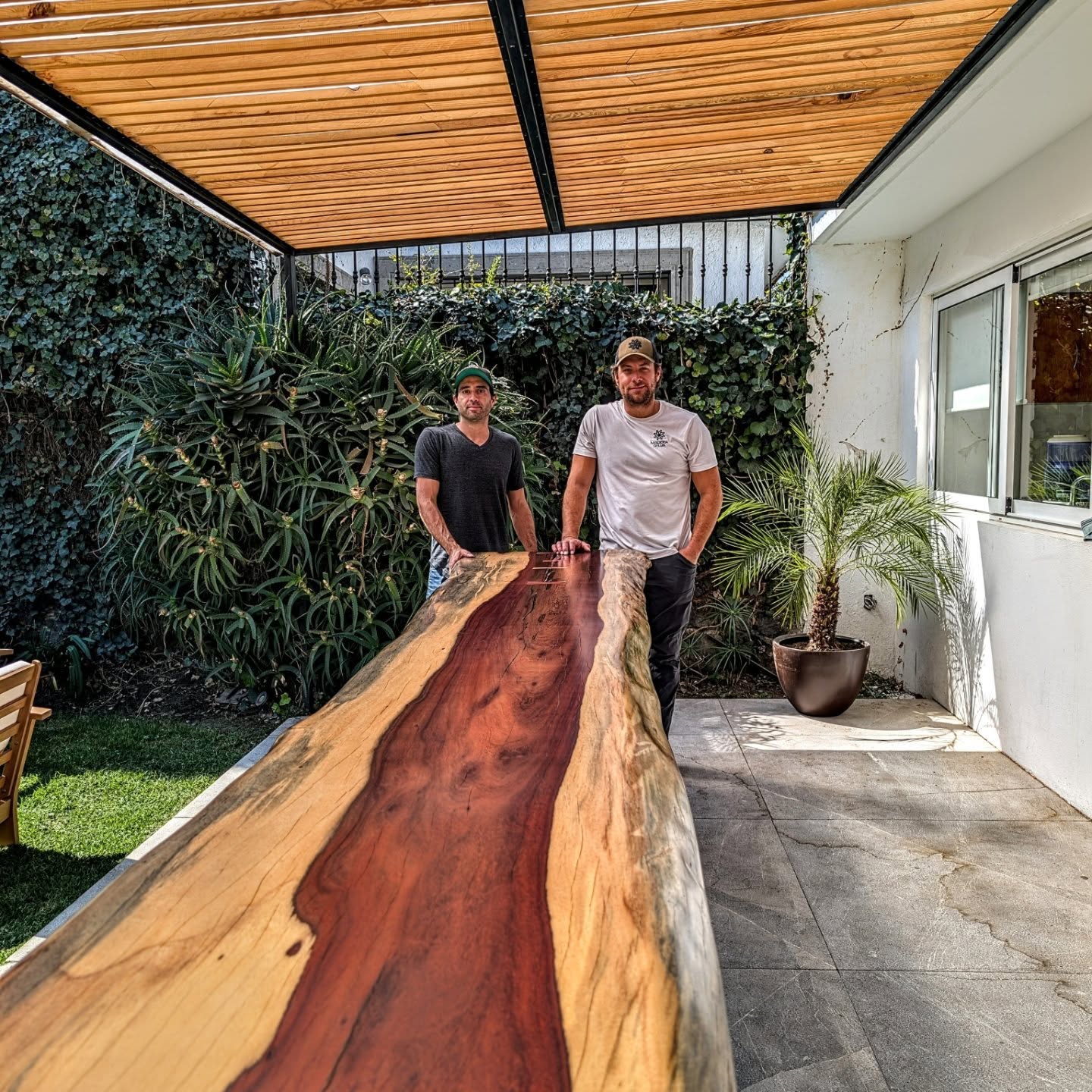 Two Madera Selva craftsmen standing beside a single continuous live-edge slab table on a terrace